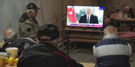Männer sitzen voreinem Fernseher, auf dem man das Gesicht von Aserbeidschans Präsidenten Alijew sieht