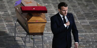 Emmanuel Macron steht mit feierlich geschlossene Augen und offenbar emotional gerührt vor einem Sarg, mit dem Rücken zum Sarg
