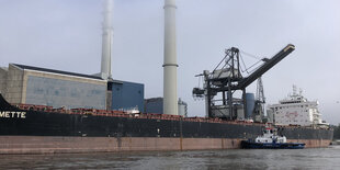 Eine großes Schiff am Kai vor einem Gebäude mit zwei langen Schornsteinen und einem Kran mit langem Arm