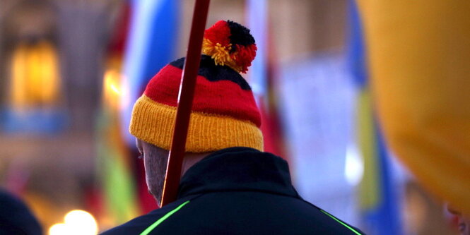 Ein Mann mit einer schwarz rot goldenen Pudelmütze ist von hinten zu sehen. Er trägt während einer Pegida Demonstration eine Fahne.