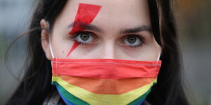 Eine Demonstrantin hart roten Pfeil uber das rechte Auge geschminkt und trägt einen regenbogenfarbenen Mund-Nasen-Schutz