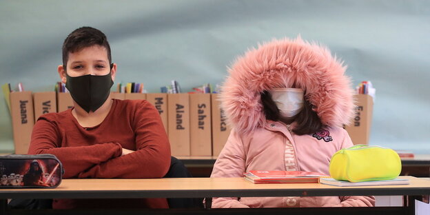 Eine Schülerin trägt eine Mundschutzmaske und hat die Fellkapuze ihres Anoraks angezogen, neben ihr sitzt ein Mitschüler mit Mundschutzmaske