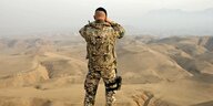 Ein Bundeswehr-Soldat ist von hinten zu sehen . Er steht auf einem Hügel und sieht mit einem Fernglas in eine afghanische Steppenlandschaft