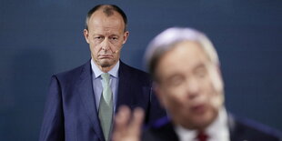 Friedrich Merz steht mit skeptischen Blick hinter Armin Laschet, der unscharif im Vordergrund des Bildes zu sehen ist.