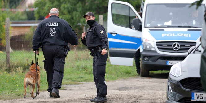 Zwei Polizisten laufen auf ein Grundstück. Sie haben einen Schäferhund dabei. Neben ihnen parkt ein Einsatzfahrzeug.