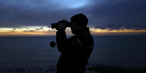Ein Mann blickt mit Fernglas kurz nach Sonnenuntergang in Richtung Meer.