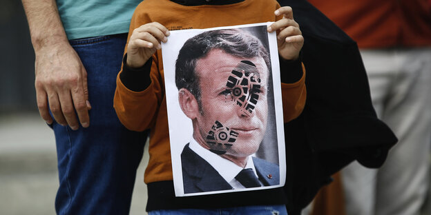 Ein Kind hält ein Plakat in den Händen auf dem Emmanuel Macron abgebildet ist in den Händen. Über dem Gesicht von Macron ist der Fußabdruck eines Stiefels zu sehen.