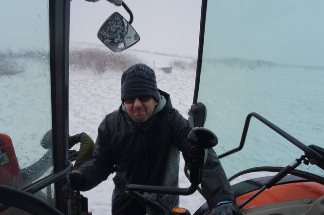 Ein Mann blickt mit zusammengekniffenen Augen in einen Traktor, hinter ihm Schnee und offenbar Wind und Kälte