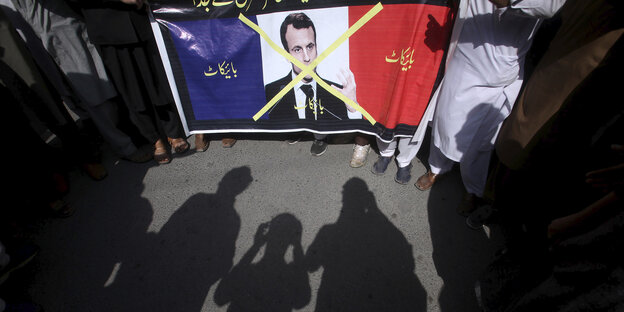 Die Silhouetten von drei Personen vor der Französischen Flagge auf der Emmanuel Macron abgebildet und durchgestrichen ist.