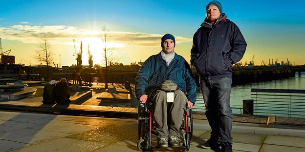 Ein Rollstuhlfahrer und ein junger Mann stehen vor der Silhouette des Hamburger Hafens.
