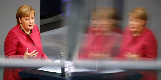 Angela Merkel trägt ein rotes Jacket und Spricht im Bundestag und spiegelt sich doppelt in einer Glasscheibe.