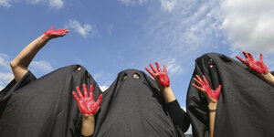 Drei Personen mit schwarzen Umhängen verkleidet heben ihre rot bemalten Hände in die Höhe