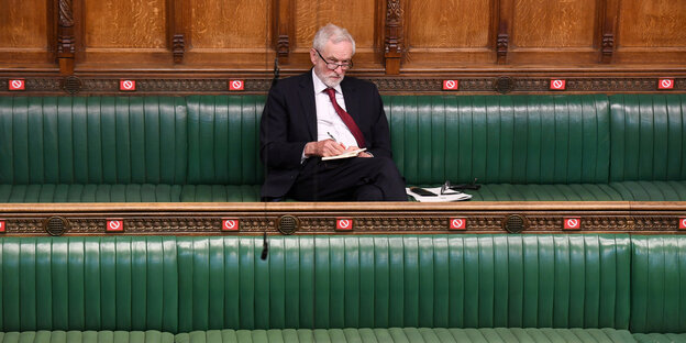 Jeremy Corbyn sitzt auf einer grünen Bank im britischen Parlamentssaal und macht sich Notizen