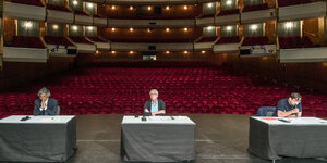 Drei Männer an weit auseinader stehenden Tischen auf der Bühne der Hamburger Staatsoper
