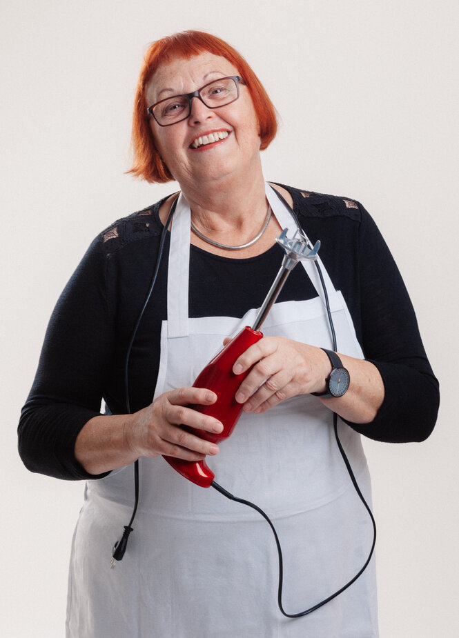 Eine lachende Frau mit Schürze, in der Hand ein roter Pürierstab