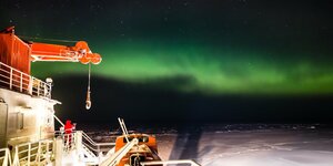 Grüne Polarlichter am Nachthimmel über dem Weddellmeer, am Bildrand eine Forschungsstation mit Krahn.