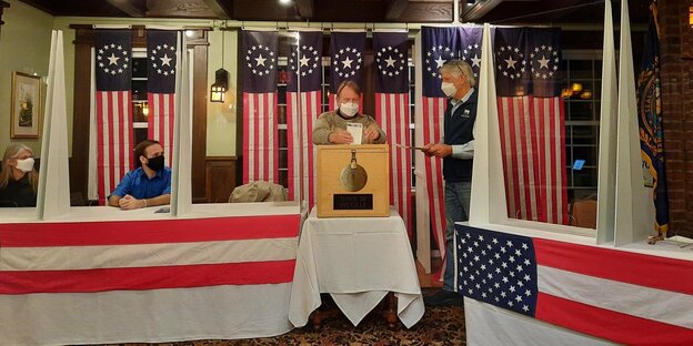 Ein Wähler steckt seine Wahlunterlagen in eine Wahlurne in Dixville Notch in den USA. Neben ihm sitzt ein Wahlhelfer, auf der anderen Seite neben ihm steht ein Wahlhelfer. Im Hintergrund und an den Tischen sind US-Flaggen befestigt.