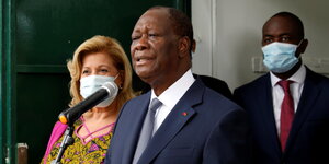 Präsident Alassane Ouattara Spricht in ein Mikrofon. Hinter ihm steht ein Mann und eine Frau mit Mund-Nasen-Bedeckung.