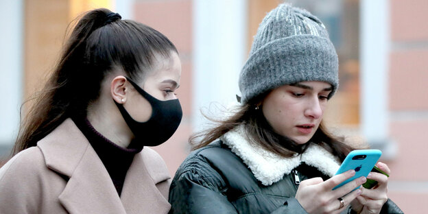 Zwei Freundinnen schauen gemeinsam auf ein Smartphone. Eine von beiden trägt einen Mund-Nasenschutz.