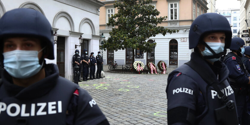 Polizisten mit Mundnasenschutz und Helmen stehen wachsam herum, hinter ihnen ist die Innenstadt zu sehen, an einer Stelle wurden Kränze hingelegt