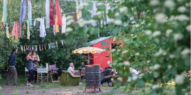 Die Subkultur-Location "Irgendwo" in Bremen an einem Sommertag. Einige junge Menschen sitzen draußen auf Sesseln, andere stehen daneben. In der Mitte ist eine Feuertonne zu sehen, im Vordergrund flattern bunte Stofffetzen von einer Leine.