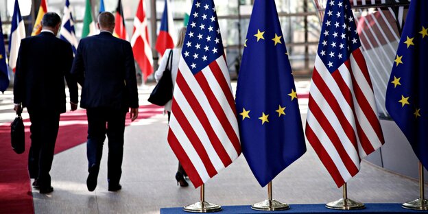 Zwei Männer in Anzügen laufen im Europäischen Rat in Brüssel neben Flaggen der USA un Europa.