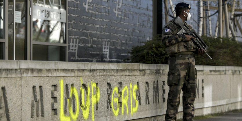 Ein bewaffneter Soldat steht einer Gedenkstätte in Lyon - Der Schriftzug Loup Gris ist mit gelber Farbe aufgesprüht.