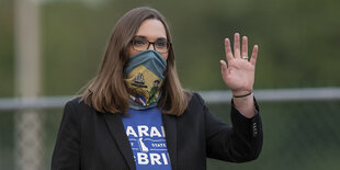 Sarah McBride mit Mund-Nasenbedeckung hebt die linke Hand zum Gruß