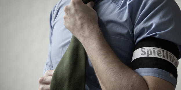Ein Mann bindet sich den Schlips. Am Arm trägt er eine Armbinde mit dem Schriftzug "Spielführer".