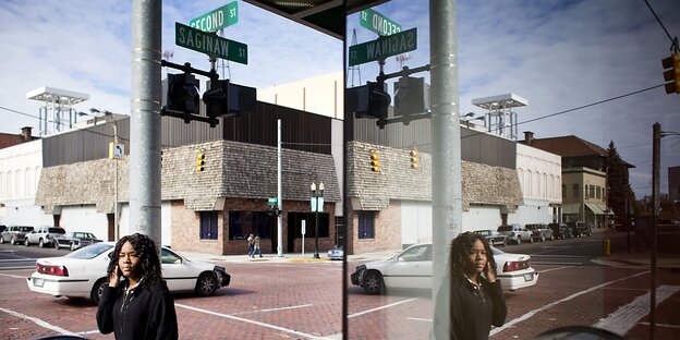 Eine schwarze Frau steht telefonierend auf einer Straße in Michigan; ihr Spiegelbild ist in einem Fenster eines Hauses zu sehen