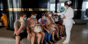 Touristen und Touristinnen mit Mundschutzmasken warten daruaf, dass eine Frau die Körpertemperatur misst