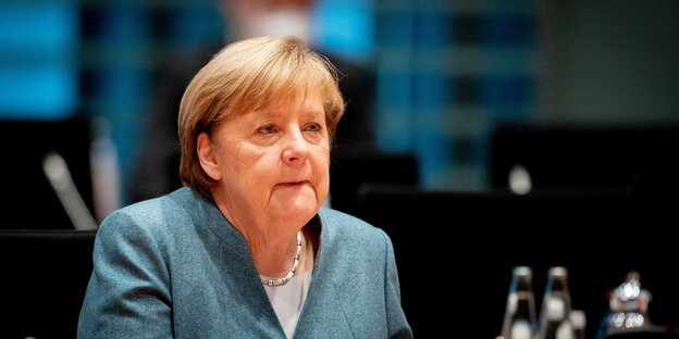 Angela Merkel sitzt in blauem Blazer und mit geschlossenem Mund vor einem Mikrofon.