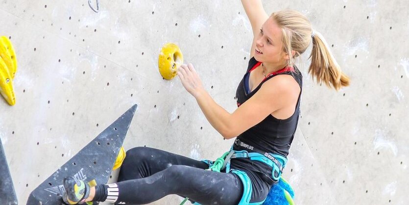 Eine junge Frau in einem Klettergeschirr hält sich mit einer Hand an einem Griff fest und greift mit der anderen nach dem nächsten