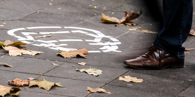 : Ein Piktogramm, das auf das Tragen einer Schutzmaske hinweist, ist auf den Bürgersteig gemalt. Ein Manng eht vorbei.