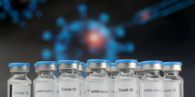 Impf-Fläschchen stehen vor einer blauen Illustration eines Coronavirus und einer Spritze