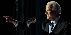Joe Biden vor schwarzem Hintergrund zeigt mit der rechten Hand nach links, während sich die Hand spiegelt und damit auf Joe Biden selbst zeigt.