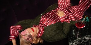 Eine Frau im gestreiften und glänzenden Pyjama liegt mit Maske über den Augen, Lippenstift und Ohrschmuck und einer grünen Jacke auf dem Boden