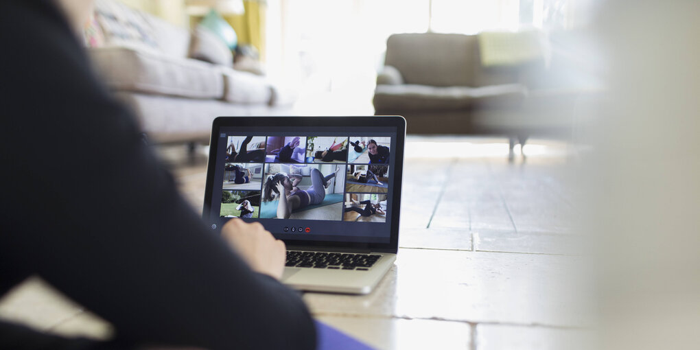 Eine Frau sitzt vor einem Laptop. Mehrere teilnehmer einer virtuellen Yoga Session sind auf dem Bildschirm zu sehen.