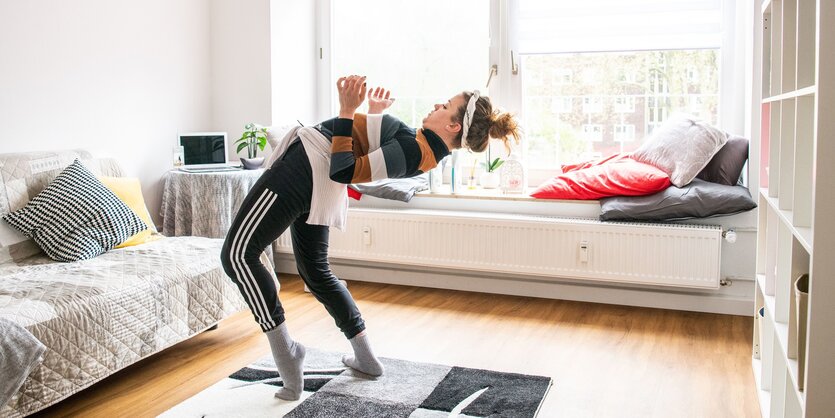 Eine Tänzerin in Jogginghose und Pulli im Wohnzimmer; sie lehnt ihren Rücken weit nach hinten in die Beuge, die Arme zeigen etwas nach oben, nur die Zehenspitzen des linken Fußes berühren den Boden