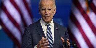 Biden steht an einem Redepult und gestikuliert, im Hintergrund US-Flaggen.