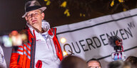 Jens Bütttner steht in Trainingsanzug und mit Hut vor einerm "Querdenken"-Banner