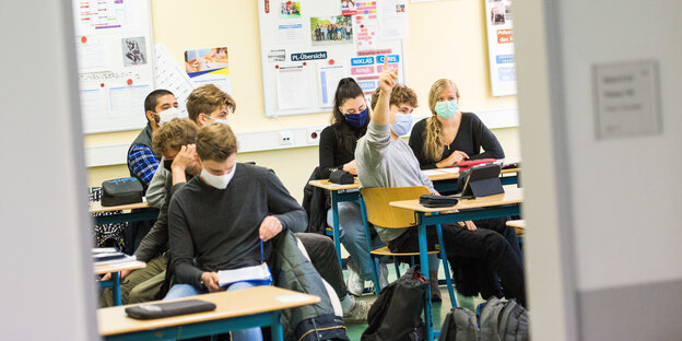 Schüler*innen mit Maske im Klassenraum, eine Schülerin im Vordergrund trägt zusätzlich eine Wollmütze