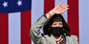 US-Vizepräsidentin Kamala Harris vor einer großen USA Flagge hebt die rechte Hand.