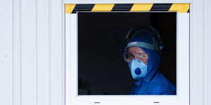 Ein Mitarbeiter mit FFP-2-Maske und blauem Schutzkittel schaut aus dem Fenster einer Corona-Test-Station in einem Container