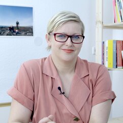 Stine Marie Javobsen, eine Frau mittleren Alters mit Brille. Sie hat ihr Haar nach hinten gebunden und trägt eine weite Bluse. Hintr ihr eine weiße Wand mit einer großen Fotografie und ein Bücherregal aus einfachen Holzbrettern.