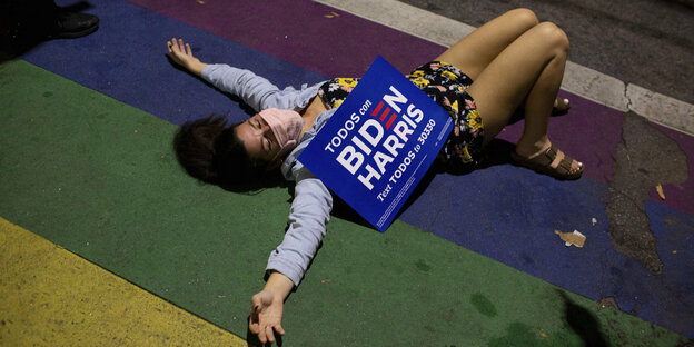 Eine Frau liegt auf dem Boden, auf ihr leigt ein Plakat mit der Aufschrift "Biden Harris"