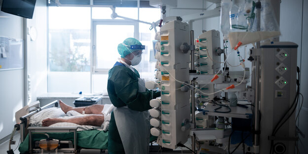 Patient*innenzimmer auf einer Intensivstation im Krankenhaus. Im Bild links steht ein Bett, zu sehen sind die nackten Beine eines Mannes. Die Krankenpflegerin mit grüner Schutzkleidung bedient eine Maschine