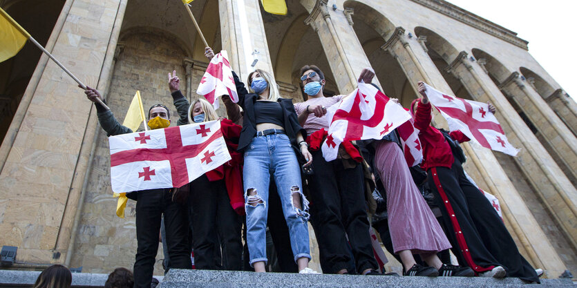 Knapp zehn Menschen demonstrieren vor dem Parlamentsgebäude in Tiflis, Georgien. Sie stehen vor Säulen und schwanken georgische Fahnen.