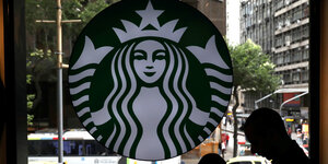 Das Logo von Starbucks an einer Fensterscheibe. Es zeigt eine Frau mit langen, welligen Haaren, die eine Krone trägt. Vor dem Fenster die Silhouette eines Mannes