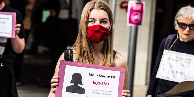 Eine Frau mit Gesichtsmaske zeigt ein Schild: mein Name ist Inge (78) ich wurde von meinem Ehemann ermordet